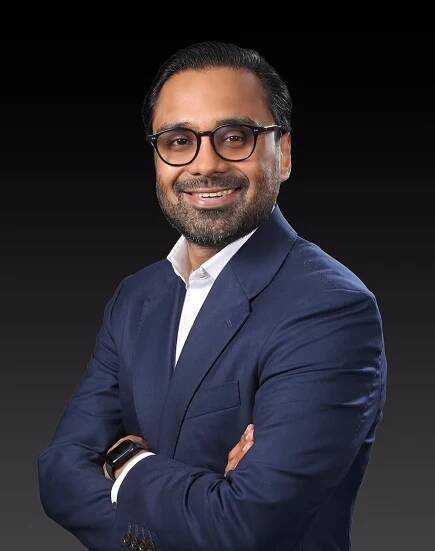Zoheb Amin Khan, Non-Executive Director, wearing glasses and a dark suit over a white shirt, arms crossed, smiling confidently.