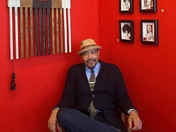 A man sits in a chair against a bright red wall adorned with a large decorative comb and a group of framed black-and-white portraits.