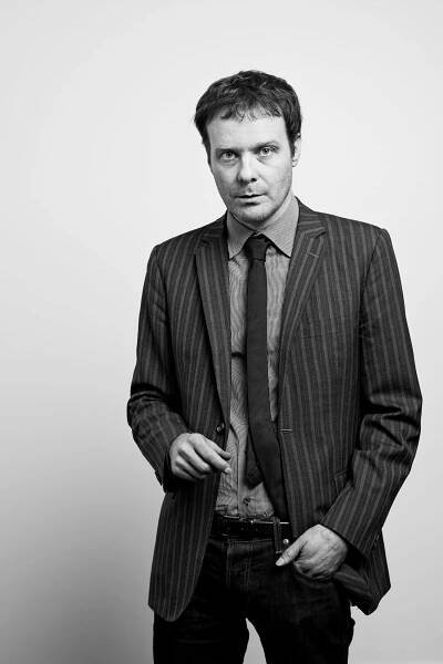 A black and white portrait photo of a White, dark-haired man in a dark striped jacket, black tie, and dark jeans. He has his right hand in his pockets.