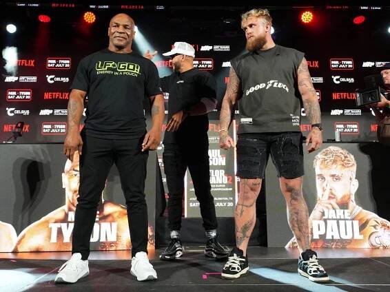 A picture of Mike Tyson and Jake Paul in casual outfits standing on a stage, with Paul staring a Tyson.