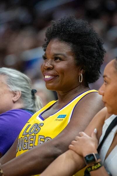 Leslie Jones smiling at a sports event, wearing a sleeveless jersey with "Los Angeles" printed. Seated among others and enjoying the event