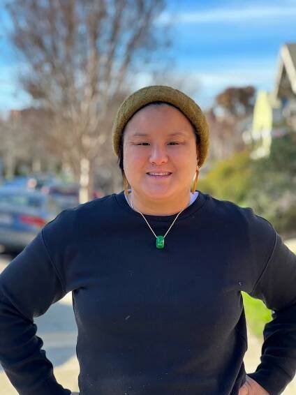 impage of person wearing a brown beanie and dark sweater with a necklace and earrings, smiling in a sunny green background