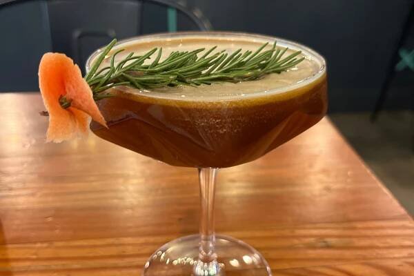 A coupe-style glass with a coffee drink in it garnished with a sprig of rosemary