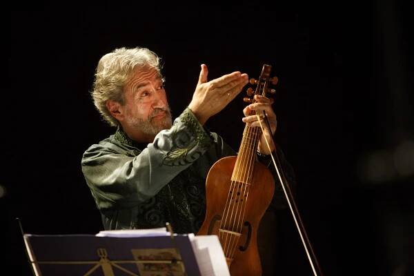 Jordi Savall, mit grauem Haar und einem grün gemusterten Hemd, hält ein hölzernes Saiteninstrument in der Hand. Er steht auf einer schwach beleuchteten Bühne. Im Vordergrund sind Notenblätter und ein Notenpult zu sehen.