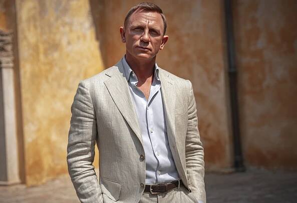 Daniel Craig in a beige linen suit and light blue shirt standing confidently against a sunlit rustic wall as James Bond