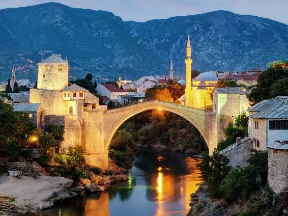 Večerní Mostar s osvětleným mostem, strážní věží a minaretem v pozadí