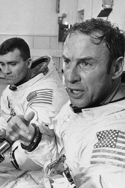 A black-and-white photo of James Lovell, wearing a white space suit decorated with an American flag, sitting next to another astronaut and gesturing with one hand.