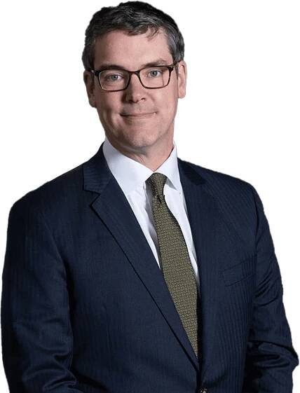 Man in a dark suit and glasses standing against a plain background.