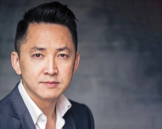 A photo of Viet Thanh Nguyen, a person with brown skin and short straight black hair. He wears a white collared shirt and a gray suit. He faces the camera.