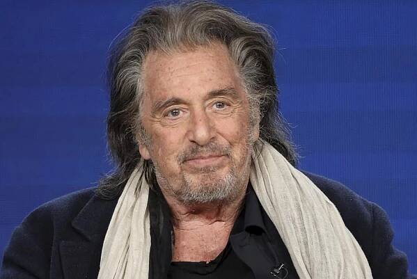 Al Pacino is wearing a black coat with a beige scarf as he smiles and sits on a stage.