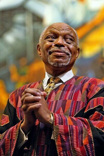 The Rev. Cecil Murray, an older man with balding gray hair and a beard wearing a patterned robe over a white-collared shirt and tie, holding his hands together while looking away from the camera.