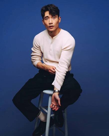 A person sits on a stool in a casual outfit with a light long-sleeve shirt and dark pants against a plain background