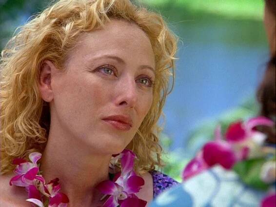 Virginia Madsen as Emma Burke in Returning Lily (AKA Tempted), wearing a floral dress and a purple orchid lei. She gazes emotionally at someone off-camera, with a lush, tropical setting in the background.