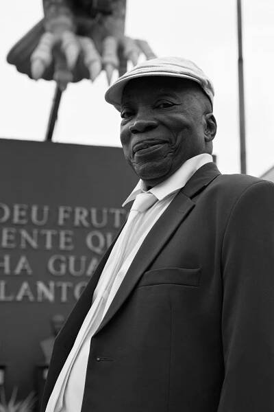 Black and white portrait of a smiling elderly man wearing a suit and flat cap standing outdoors.