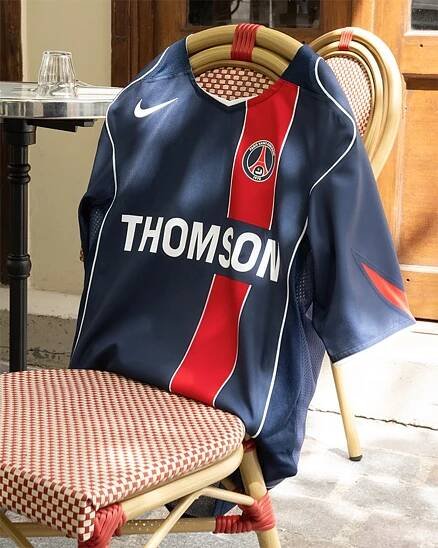 Classic Paris Saint-Germain home football jersey in navy blue with a red vertical stripe, Nike logo, and Thomson sponsor, draped over a café chair in an outdoor Parisian setting.          Spørg ChatGPT