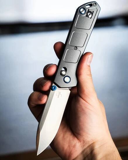 Hand holding a folding knife with a silver blade and a gray handle featuring blue accents and screws. The blade has a sharp, pointed tip and a clean, polished finish. The handle has a textured surface with rectangular indents and a thumb stud for opening.