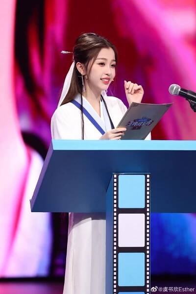 a woman standing at a podium with a book in front of her and holding a microphone