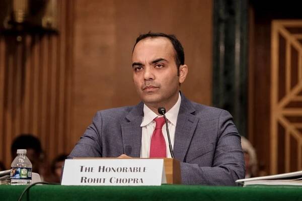 Rohit Chopra, in a suit and a red tie, sitting at a table with a name placard and a microphone in front of him.