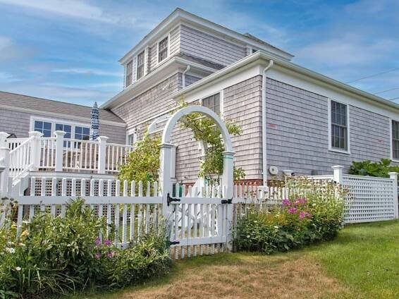 A white picket fence with an arched gate surrounds a garden in front of a gray shingle-style house with a raised deck and an umbrella. The sky is partly cloudy.