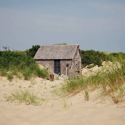 A wooden shingled-covered shack sits among sand and grass on Cape Cod