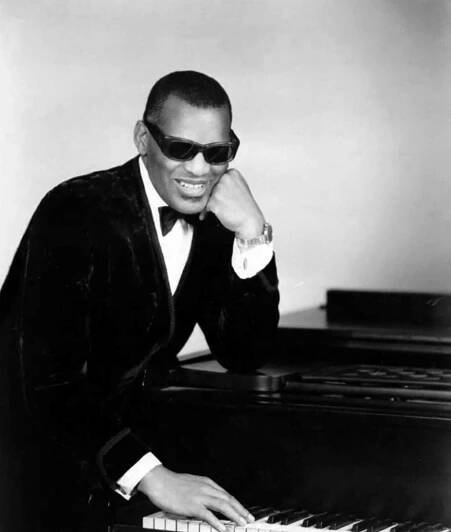 A smiling Black man wearing a tuxedo and sunglasses, has one hand on his face, the other on piano keys.