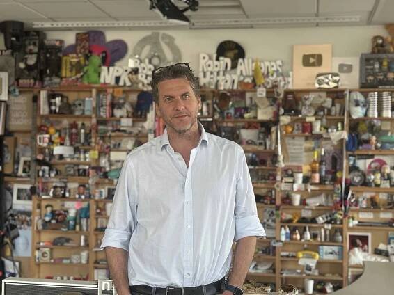 Tommy Evans in a button down shirt with the sleeves rolled up and his hands in his pocket looks at the camera from NPR's Tiny Desk studio. 