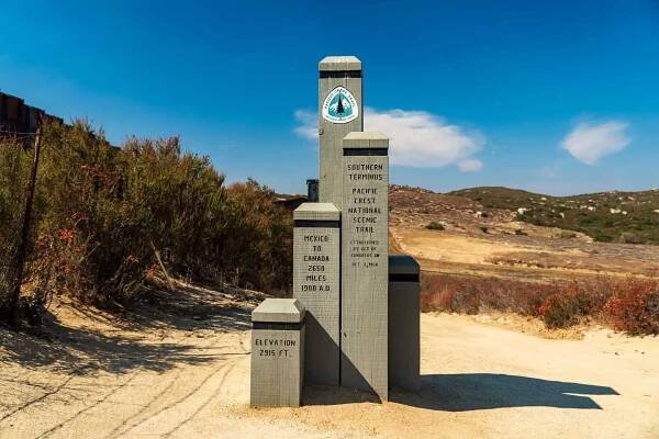 A stone monument marks the southern terminus of the Pacific Crest National Scenic Trail, with inscriptions detailing the trails mileage and elevation. Surrounded by dry vegetation and hills under a clear blue sky, it stands ready for those embarking on upcoming pct surveys in 2024.