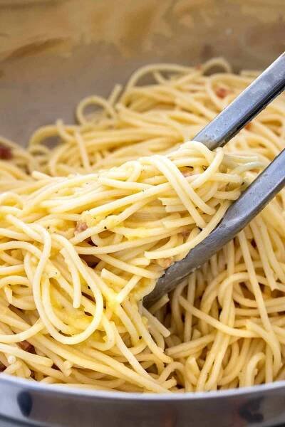 Metal tongs tossing spaghetti in a bowl with eggs and cheese.
