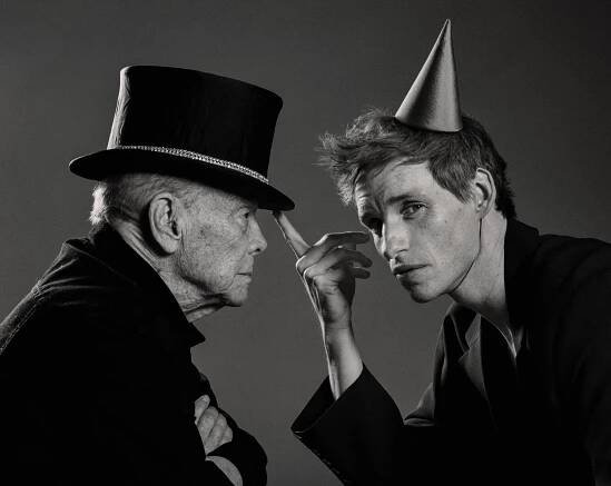 In a black-and-white photo, two men, both dressed in black, have positioned their bodies so they are facing each other. The man on the left is wearing a top hat and looking at the other man, and the other man is wearing a birthday party hat, with his head turned to look at the camera.