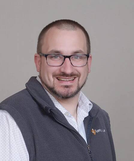 A man with short hair, glasses, and a beard smiles at the camera. He is wearing a gray fleece vest over a white shirt and standing against a plain light background, representing the friendly spirit of our team.