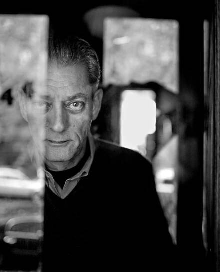 A black and white close-up photo of Mr. Auster standing to the left side of the frame looking straight at the camera, his face partly obscured behind a blurred reflection in the foreground. His hair, partly gray, is combed back, and he wears a dark V-necked sweater over a open-collared shirt.