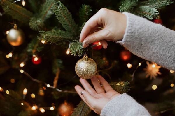 Žena zdobí vánoční stromek s lesklým zlatým ozdobou zblízka. Příprava na vánoční čas. Moderní třpytivý ornament v rukou na pozadí slavnostního stromu ve světlech. Hezké Svátky stock fotka