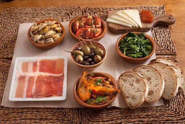 A selection of Spanish tapas including clams, ham, olives, grilled vegetables, cheese, and bread.