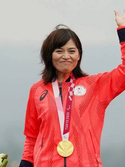 A Japanese female athlete with a gold medal around her neck waves her hand.