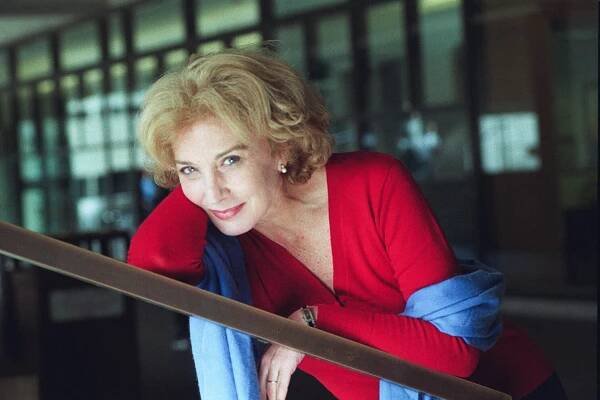 A close-up photo of her leaning against a railing and looking at the camera with a half smile. She has shoulder-length blond hair and wears a bright red sweater and blue scarf.