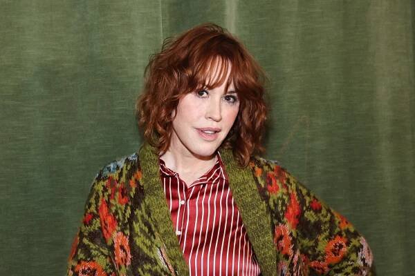 Molly Ringwald with red curly hair in a striped shirt and floral-patterned cardigan, standing against a solid backdrop