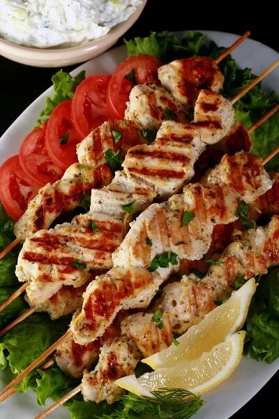 Several skewers of Green chicken souvlaki on a bed of lettuce, with tomatoes and lemon slices.