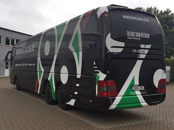 Rückansicht des Mannschaftsbusses von Hannover 96, gestaltet in Schwarz mit großen grünen und weißen grafischen Elementen. Der Schriftzug 'Niemals allein!' ist auf der Seite des Busses zu lesen. Im Hintergrund stehen Bäume und parkende Autos.