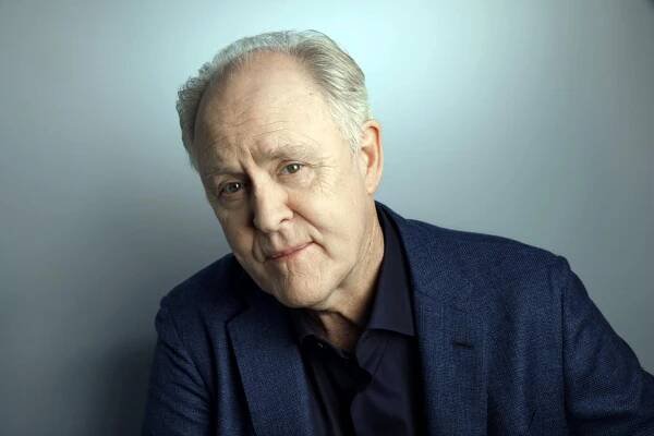 John Lithgow in a dark jacket and dark dress shirt leaning to his right and posing against a gray backdrop