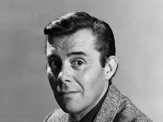 A head-shot of Dirk Bogarde staring straight at the camera. His dark hair is slicked back, he has raised his left eyebrow and he is wearing a tweed jacket.