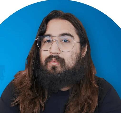 A person with long hair and a beard is wearing transparent glasses and a black shirt, standing against a solid blue background.