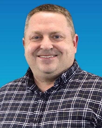 A man in a plaid shirt is smiling in front of a blue background.