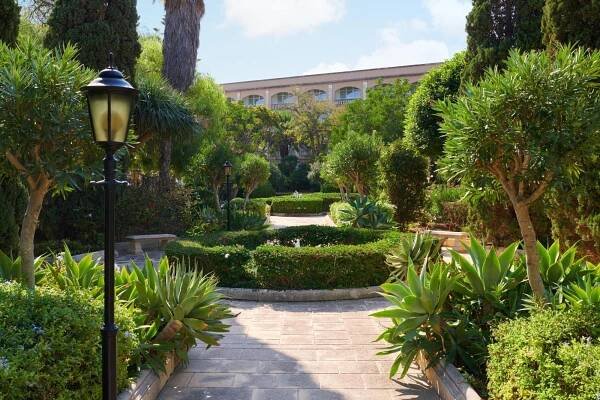 Corinthia Palace Malta Attard Exterior photo The photo shows a beautiful, lush garden with a winding stone pathway leading through it. The pathway is made of light-colored stone slabs and curves gently, inviting the viewer to explore the greenery. 

On either side of the path, there are neatly 