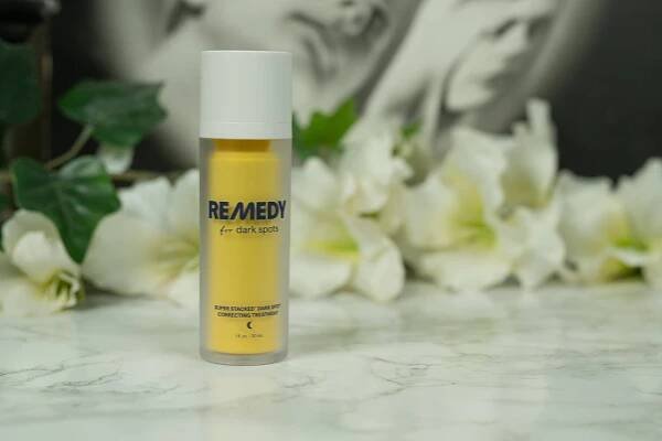 A yellow bottle of Remedy for Dark Spots standing in front of a dark background with white flowers