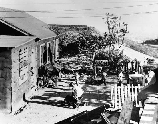 A black and white image of a ramshackle home on a hillside with people in the front lawn.