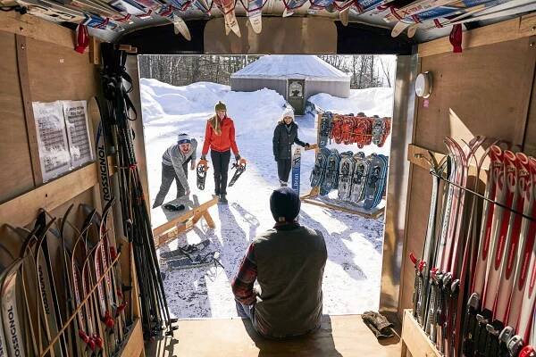 A group prepares for snowshoeing outside a cabin, surrounded by snow. Various skis and snowshoes are displayed inside the wooden cabin.