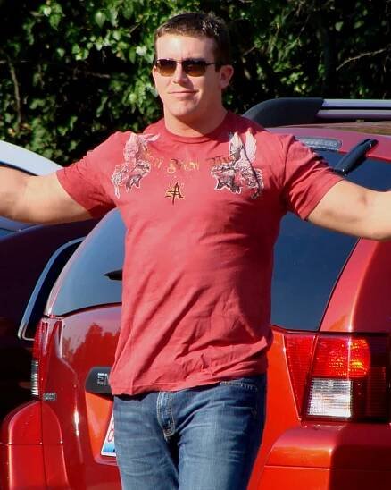 A dark haired young man wearing dark sunglasses poses for cameras with his arms outstretched. He is wearing a red T-shirt, with a pattern on both shoulders and the top part of the chest, and jeans.