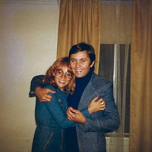 a man and woman hugging each other in front of a window with curtains behind them