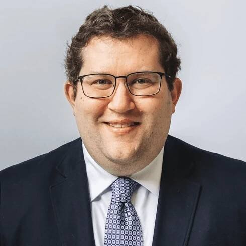 A headshot of Jeremy Young smiling at the camera, wearing a suit and glasses.