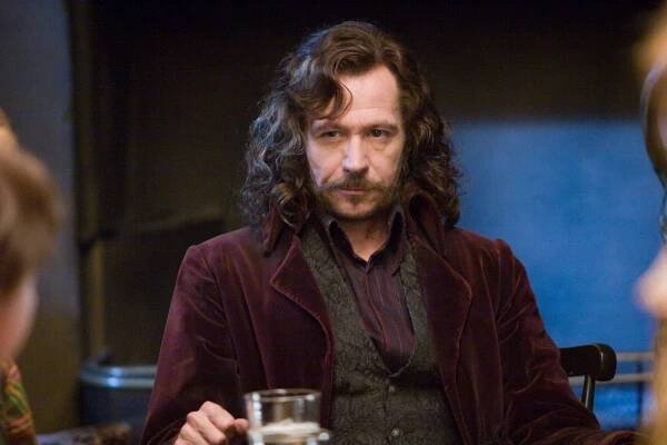 A person with long hair and a mustache, wearing a velvet jacket, sits at a table holding a glass, in what appears to be a film scene